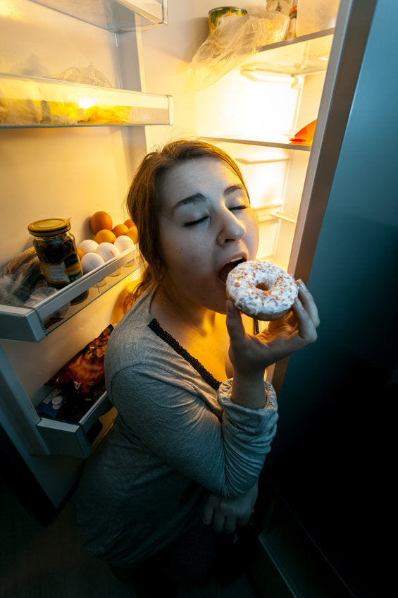 young woman with an open refrigerated door eating a glazed donut late at night